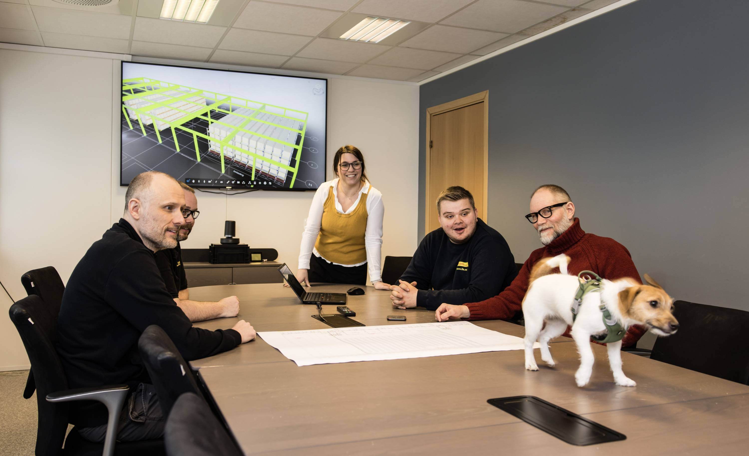 LIS:Fem personer i et møte rundt et konferansebord, med tekniske tegninger på skjermen bak og en hund som står på bordet.