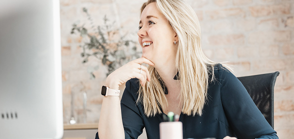 En kvinne med blondt hår sitter ved en dataskjerm og gestikulerer mens hun snakker, i et moderne kontormiljø. Bildet er tatt hos Inbound, et markedsføringsbyrå som spesialiserer seg på B2B-kunder