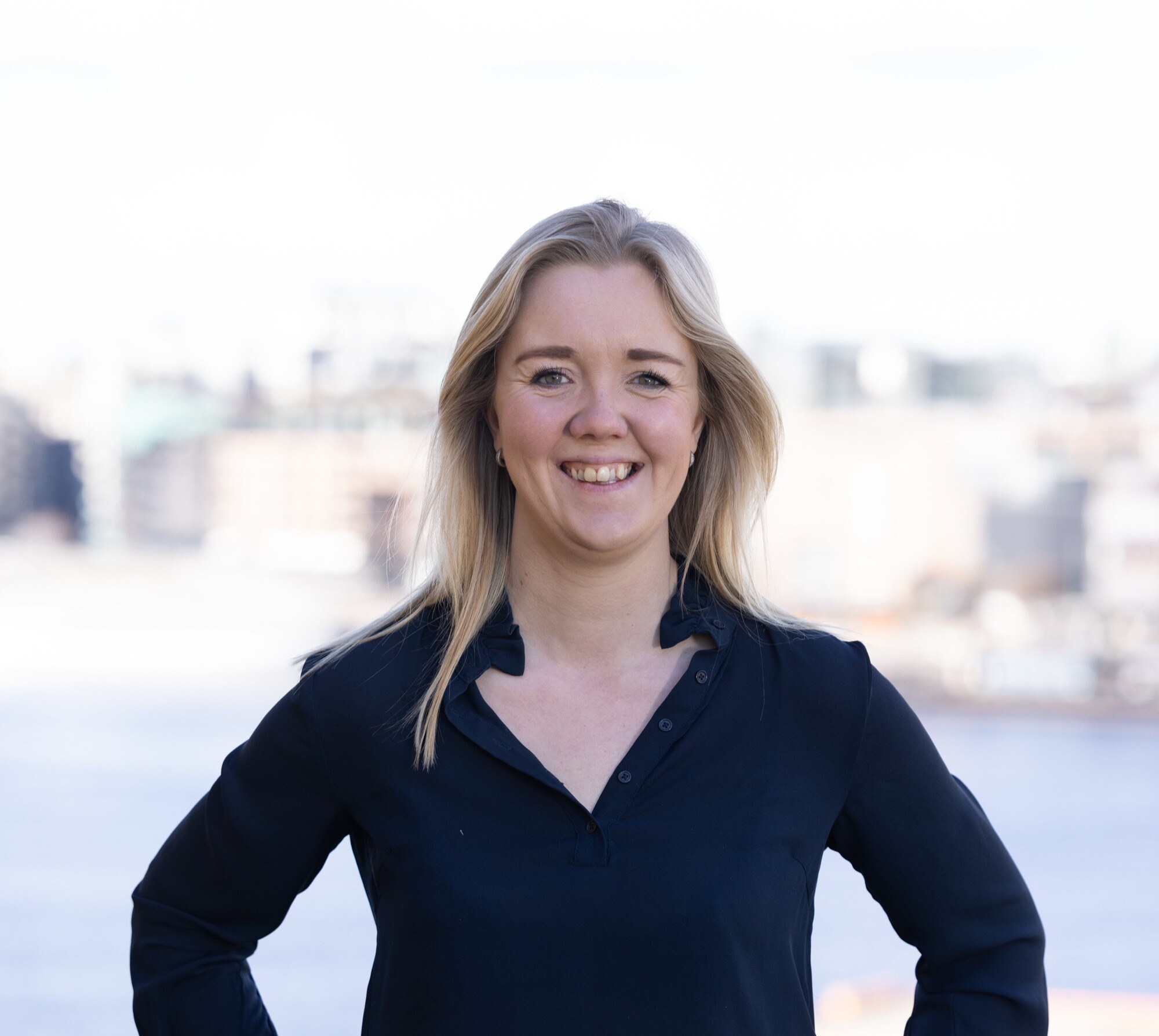 Smilende kvinne med blondt hår iført en mørkeblå skjorte, med en byutsikt i bakgrunnen.