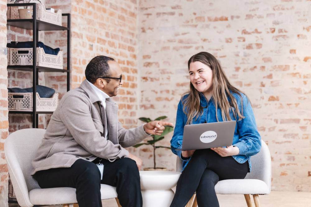En mann og en kvinne sitter i en uformell samtale i et moderne kontormiljø. Kvinnen holder en bærbar PC med Inbound-logoen, et markedsføringsbyrå som jobber med B2B-kunder.