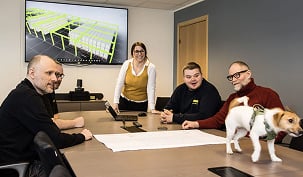 Ansatte i møte rundt et bord med plantegning på skjermen, mens en liten hund går på bordet.